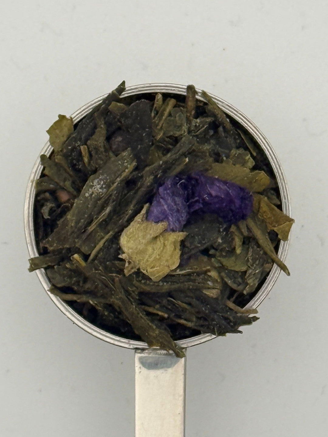 Tea leaves including green and purple elements on a metal spoon against a light gray background